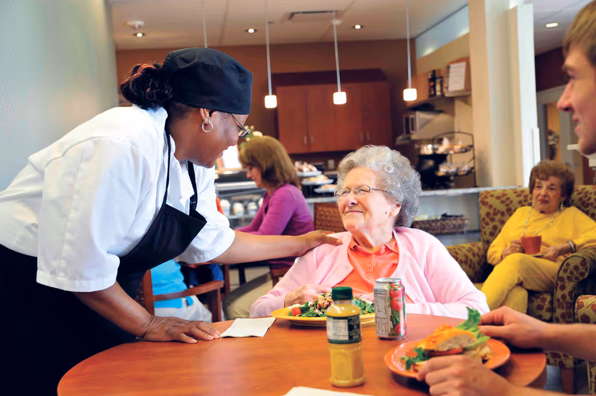 A smiling elderly woman in a pink cardigan sits at a table with a plate of food and drinks while a staff member in a white shirt and black apron leans over to talk to her. Other people are seated in the background in a cozy dining or common area with warm lighting.