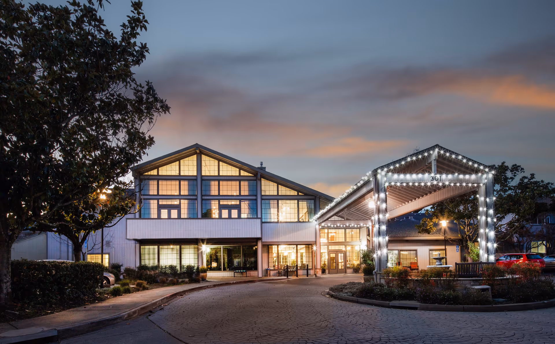 Exterior view of Oakmont Gardens senior living facility at dusk, featuring a well-lit entrance with decorative string lights outlining the covered driveway, large windows, and surrounding trees and landscaping.