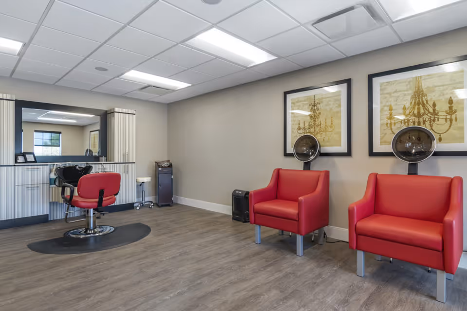 Interior of a senior living community salon area featuring a red salon chair in front of a large mirror and counter with hair care tools. Two red armchairs with hair drying hoods are positioned against the wall, which is decorated with framed artwork of chandeliers. The room has wood flooring and a drop ceiling with fluorescent lighting.