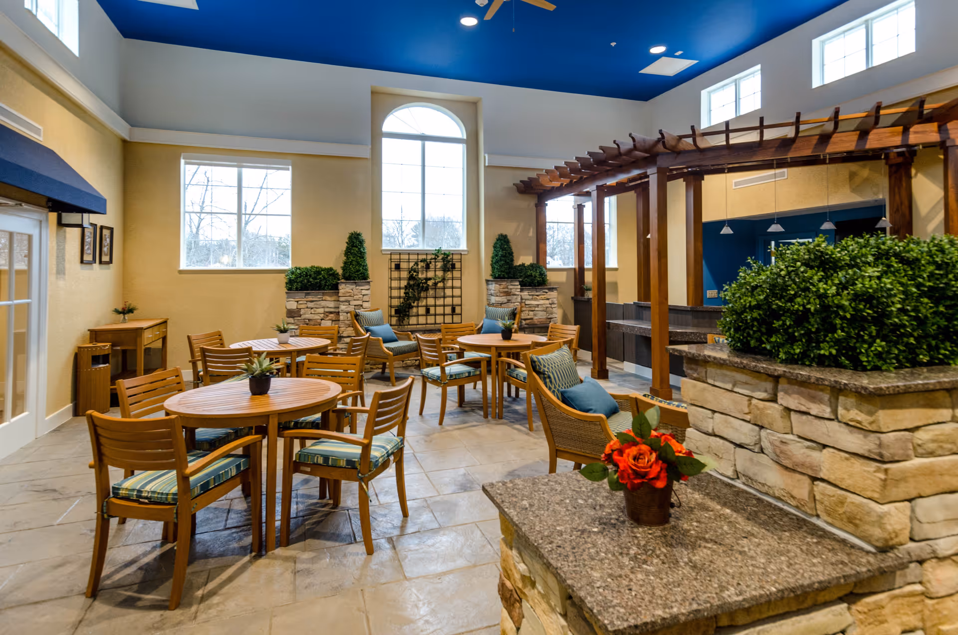 Indoor seating area with wooden tables and chairs featuring blue and green cushions, stone planters with greenery, a wooden pergola structure, large windows letting in natural light, and a blue ceiling with ceiling fans.