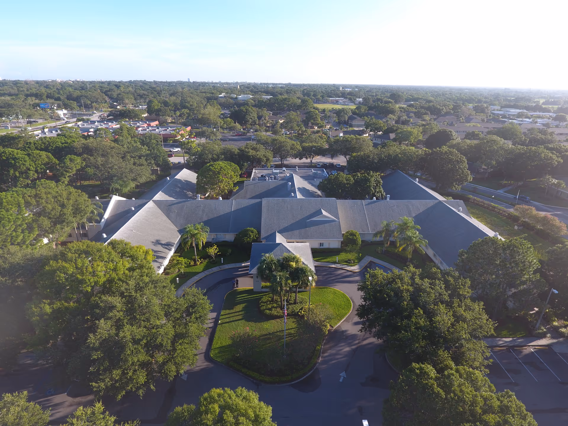 Aerial view of Palm Garden of Pinellas facility surrounded by trees and greenery, with a circular driveway and parking areas visible. The building has a large, multi-wing roof structure and is situated in a suburban area with roads and other buildings in the background.