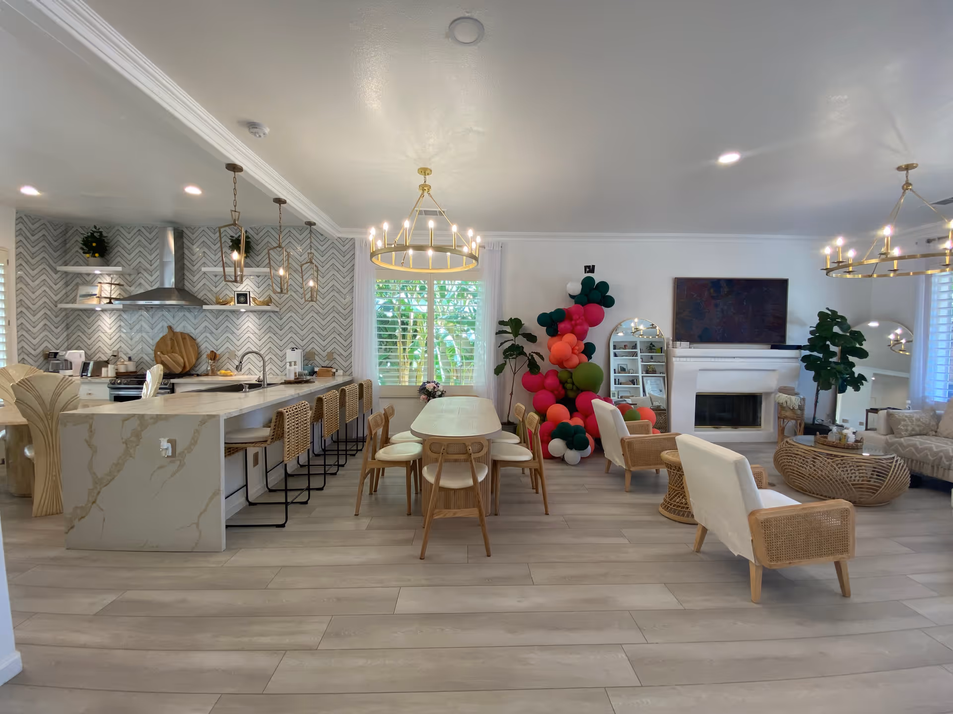 A bright and spacious open-concept living area featuring a modern kitchen with a marble island and bar stools on the left, a wooden dining table with chairs in the center, and a cozy living room with armchairs, a sofa, a fireplace, and a large TV on the right. The room is decorated with plants, colorful balloons near a window, and elegant chandeliers hanging from the ceiling.