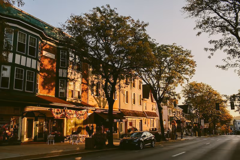 A street view of a small town with buildings featuring storefronts and apartments above. Trees line the sidewalk, and a car is parked on the street. The scene is bathed in warm, late afternoon sunlight.