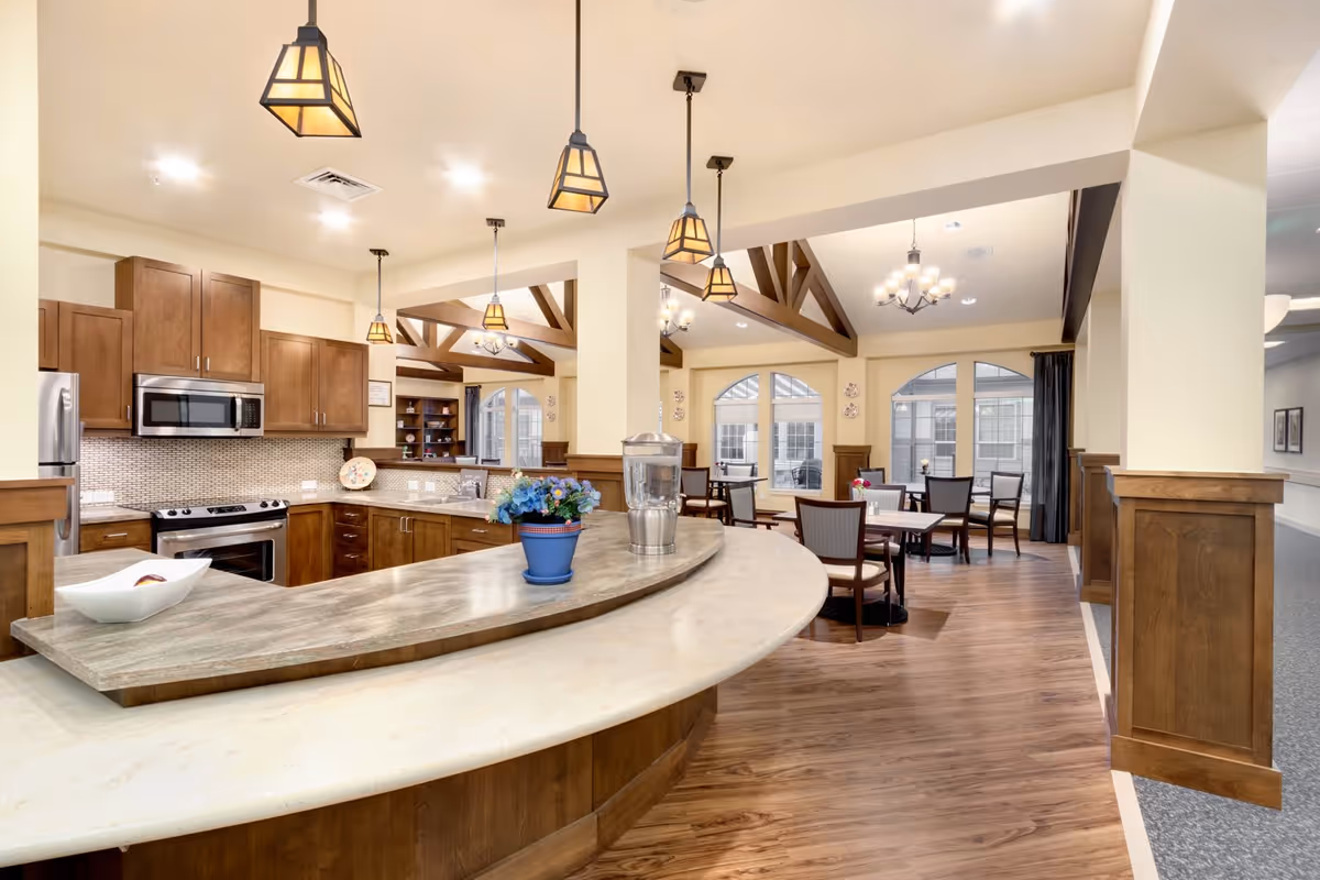 A bright and spacious kitchen and dining area in a senior living facility. The kitchen features wooden cabinets, stainless steel appliances including a microwave and stove, and a curved countertop with a decorative flower pot and water dispenser. The dining area has several tables and chairs, wooden beams on the ceiling, chandeliers, and large windows letting in natural light.