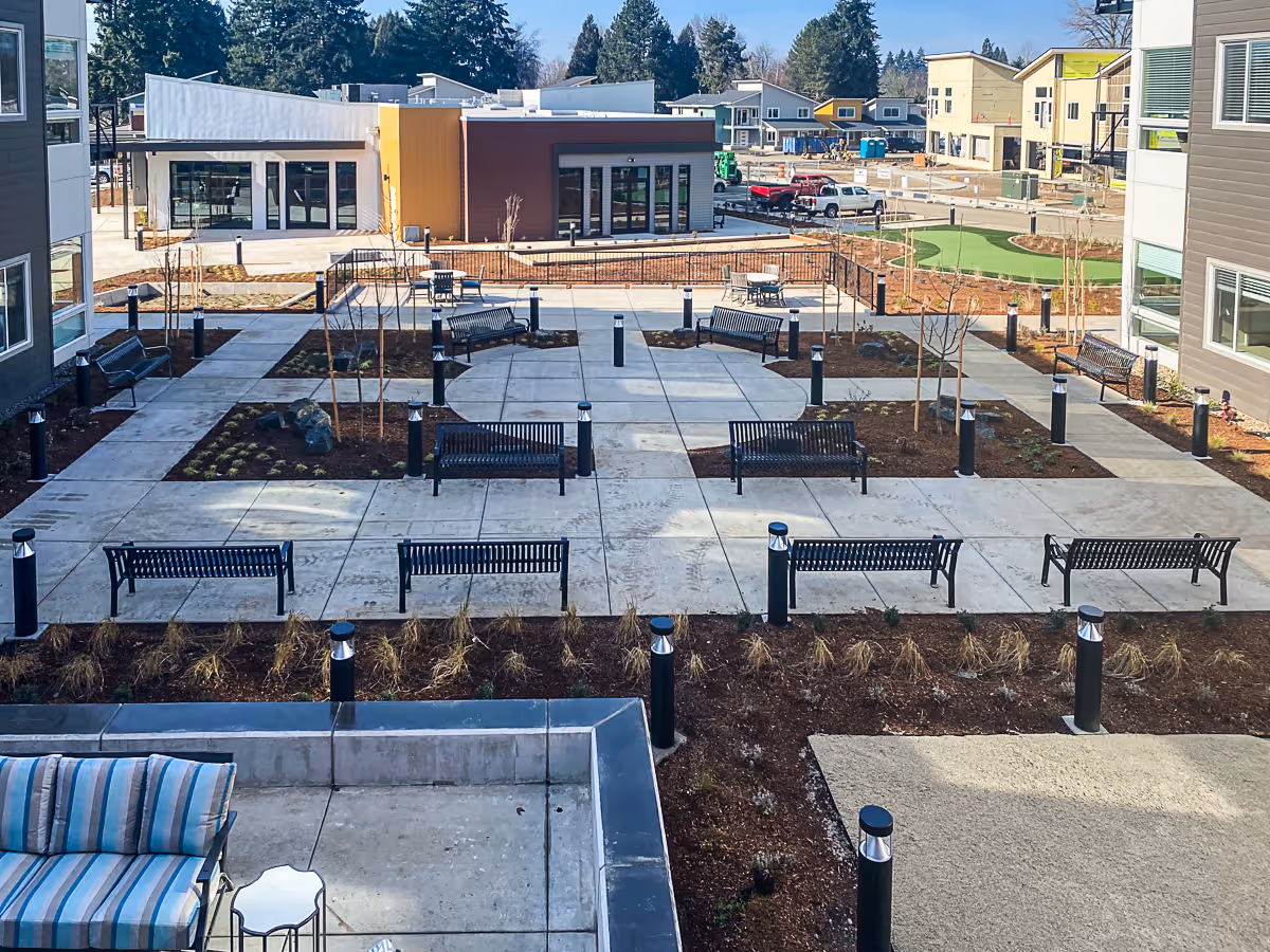 Outdoor courtyard area at Marquis Eugene Assisted Living featuring multiple black benches arranged on concrete walkways, small landscaped garden beds with young trees and plants, modern black lamp posts, and surrounding buildings in the background under a clear sky.