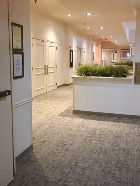 Interior hallway of a senior living facility with beige walls and carpeted floor. The hallway features double doors on the left side, a white half-wall planter with green plants, recessed ceiling lights, and framed notices on the walls.