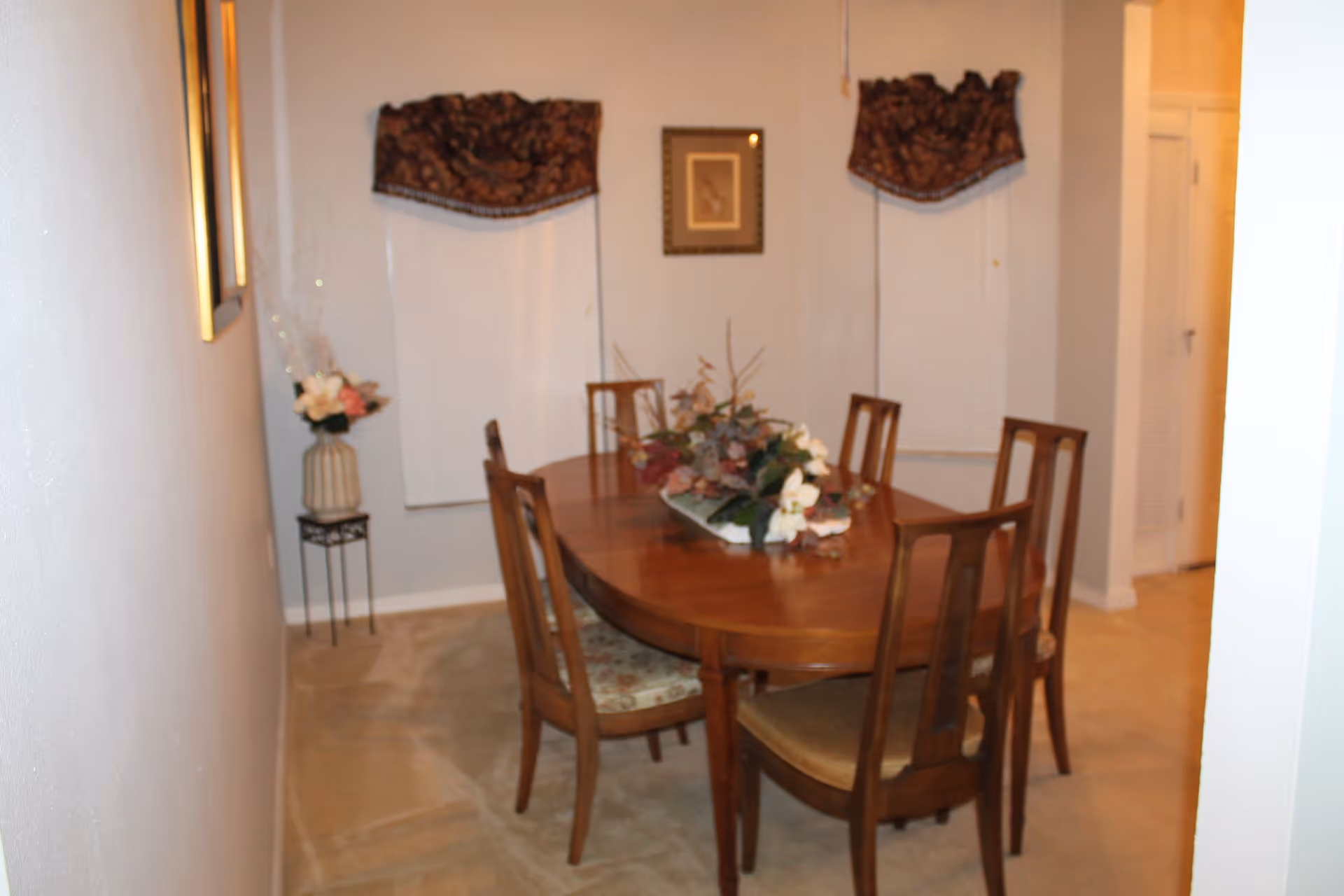 A dining room with a wooden oval dining table surrounded by six wooden chairs with cushioned seats. The table has a floral centerpiece. Two windows with dark patterned valances and white blinds are on the far wall, with a framed picture hanging between them. A small decorative stand with a vase of flowers is placed against the left wall.