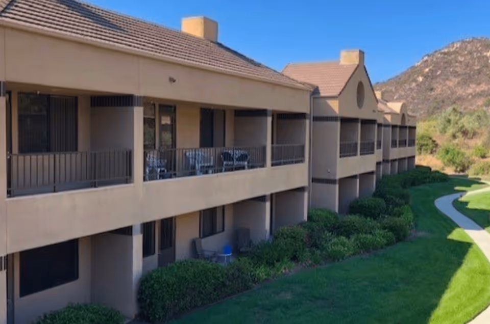 Two-story beige apartment-style building with covered balconies overlooking landscaped lawns and a curved walkway with hills in the background.