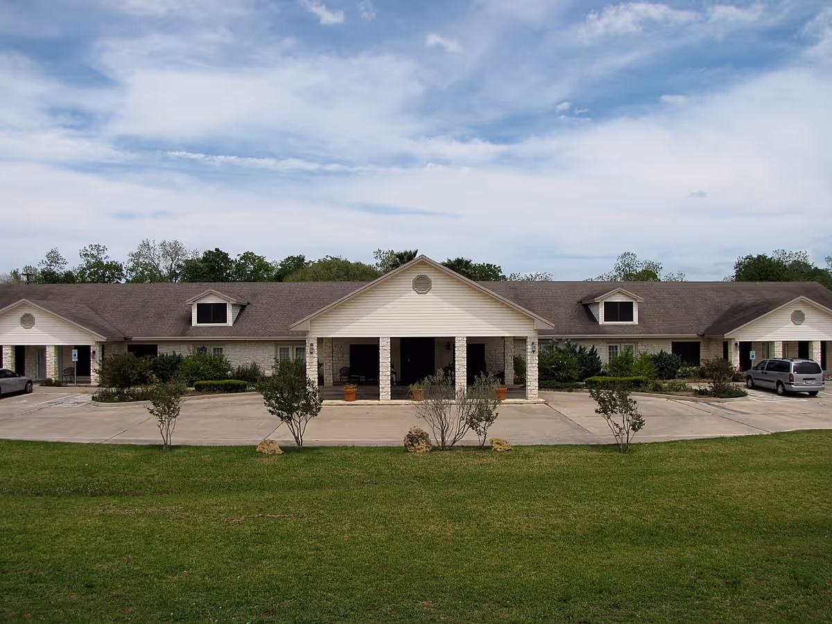 Front exterior view of a single-story assisted living facility building with a covered entrance, surrounded by a parking area and green lawn under a partly cloudy sky.