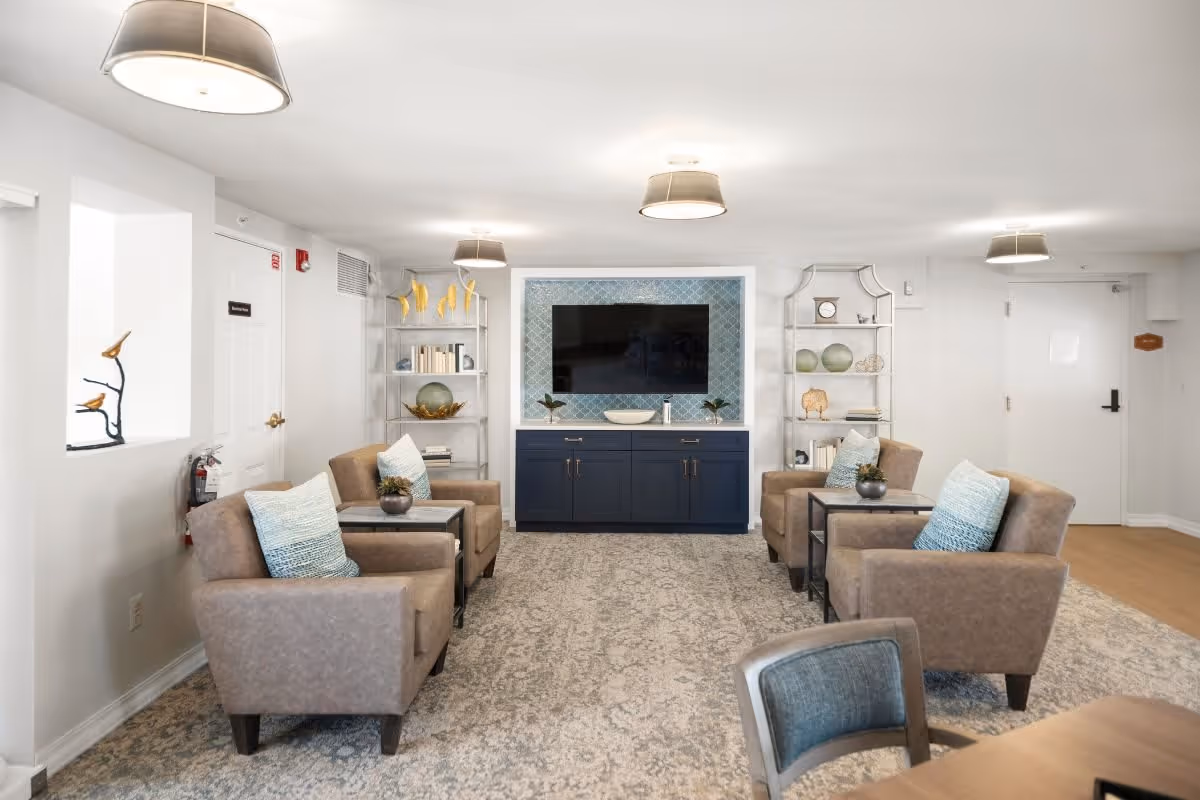 A bright and modern living room area in Pine Ridge Villas of Shelby featuring four beige armchairs with blue and white accent pillows arranged around two small side tables. The room has a large patterned rug, a flat-screen TV mounted on a blue cabinet with decorative shelving on either side holding books, plants, and ornaments. The walls and ceiling are white with three ceiling lights providing illumination.