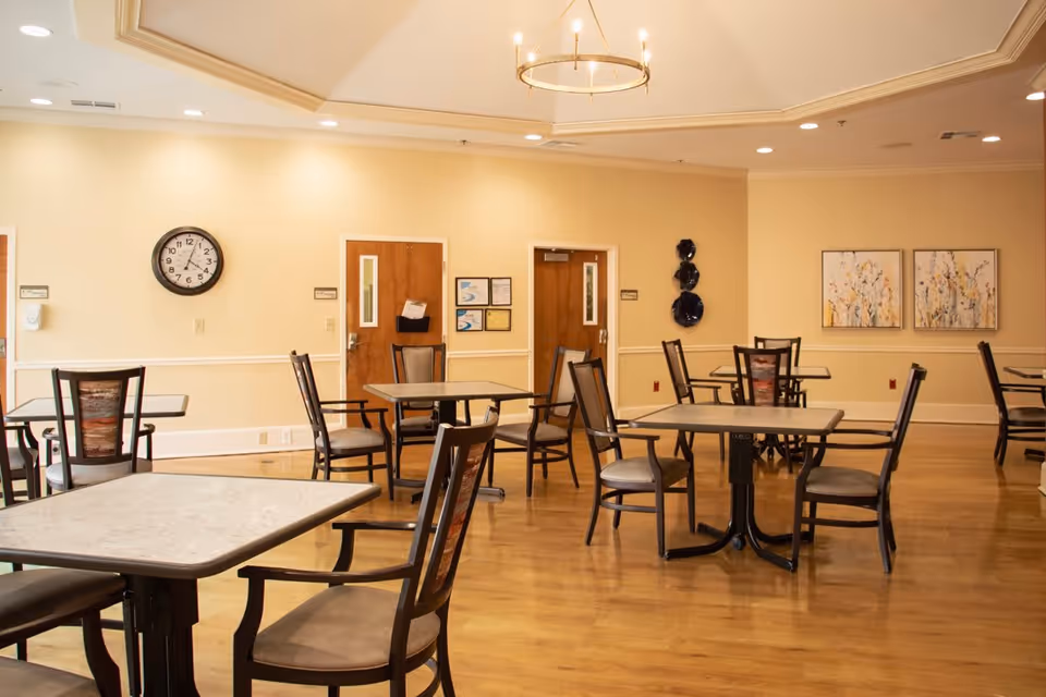 A spacious dining room with several square tables and wooden chairs with cushioned seats. The room has light yellow walls, wooden doors, a wall clock, decorative plates on one wall, and two framed floral paintings. The floor is wooden, and a chandelier hangs from the ceiling.
