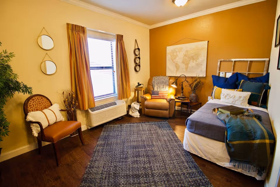 A cozy bedroom with a single bed adorned with multiple pillows and a throw blanket. Next to the bed is a small side table with a lamp and decorative items. A comfortable armchair with a patriotic pillow sits in the corner near a window with yellow curtains. A wooden chair with a cushion is placed against the opposite wall, which also features two round mirrors. The room has warm yellow and mustard-colored walls and a blue area rug on the wooden floor.