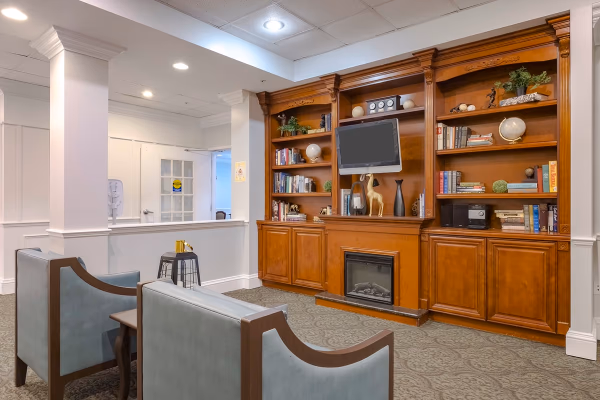 A cozy senior living common area featuring two blue upholstered chairs facing a wooden built-in bookshelf with decorative items, books, a mounted TV, and a small fireplace. The room has white walls, carpeted floor, and recessed ceiling lights.