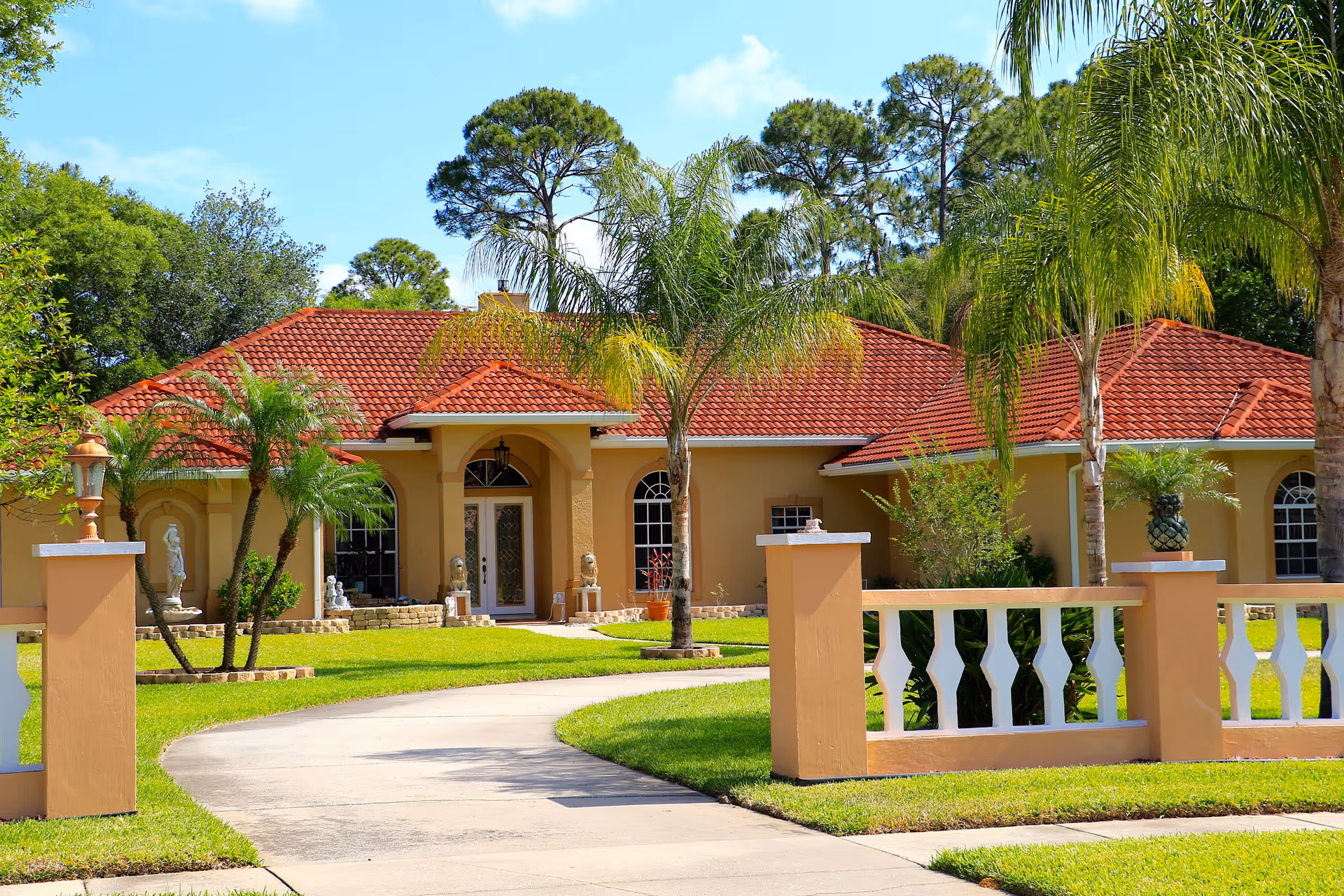 Front exterior view of a single-story house with a red tiled roof, beige walls, and a curved driveway. The yard is landscaped with green grass, palm trees, and decorative statues near the entrance.