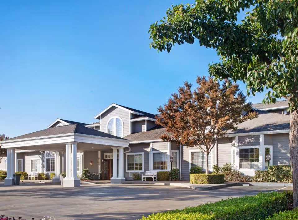 Exterior view of North Shore Place senior living facility showing a large covered entrance with white columns, multiple windows, trees with autumn foliage, and a clear blue sky.