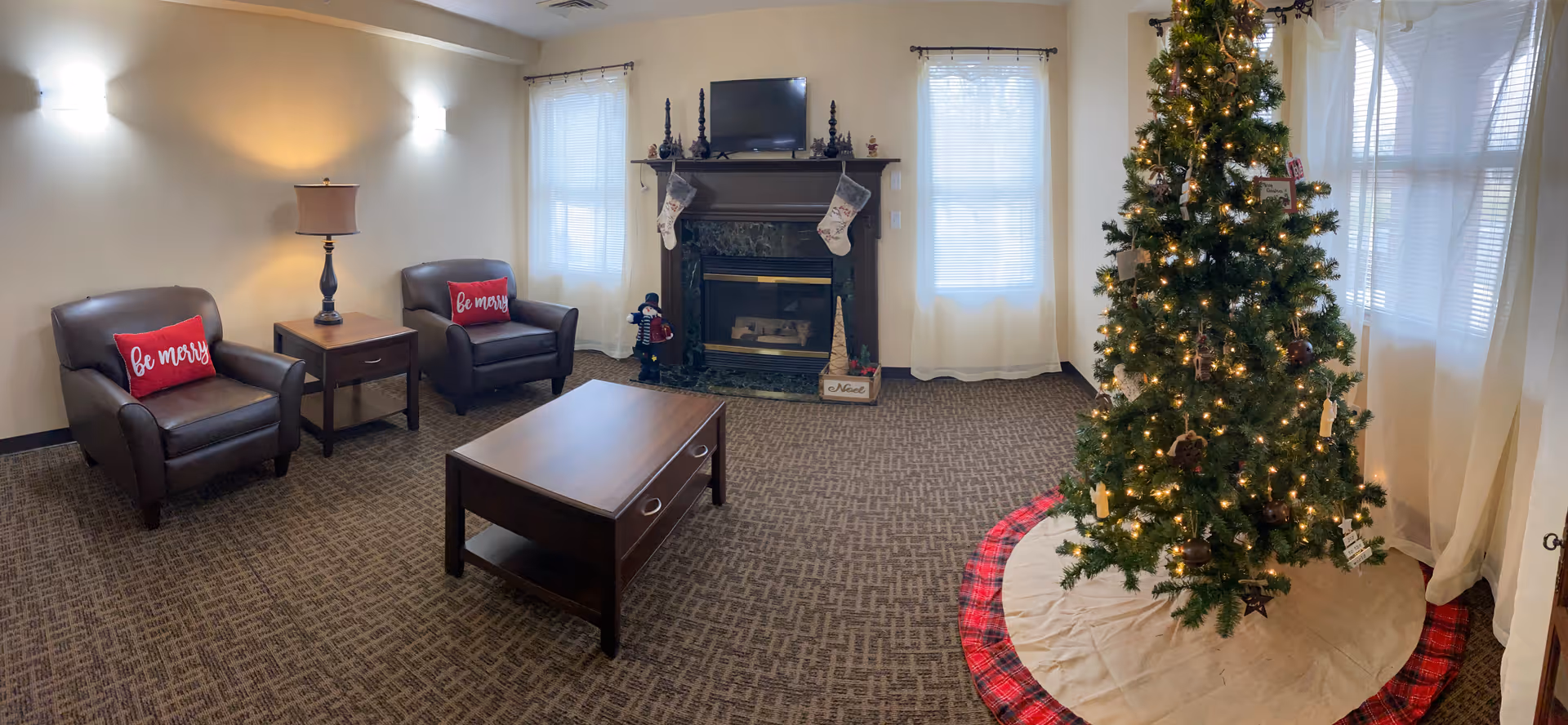 A cozy common living room with two armchairs, a coffee table, a fireplace with stockings, and a lit Christmas tree.