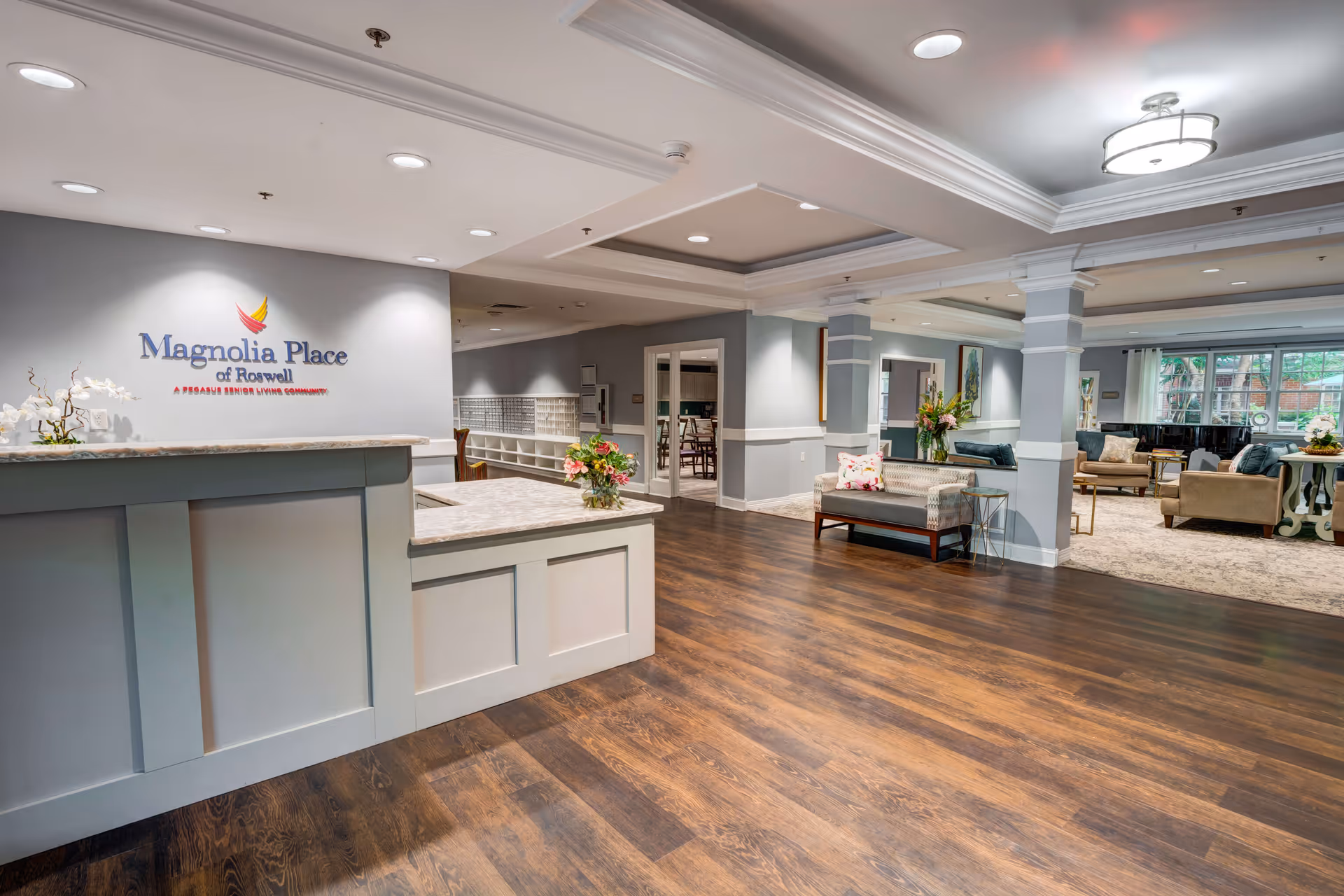 Reception area of Magnolia Place of Roswell featuring a white reception desk with a marble countertop, a sign on the wall with the facility's name and logo, wood flooring, and a spacious seating area with sofas and chairs in the background.