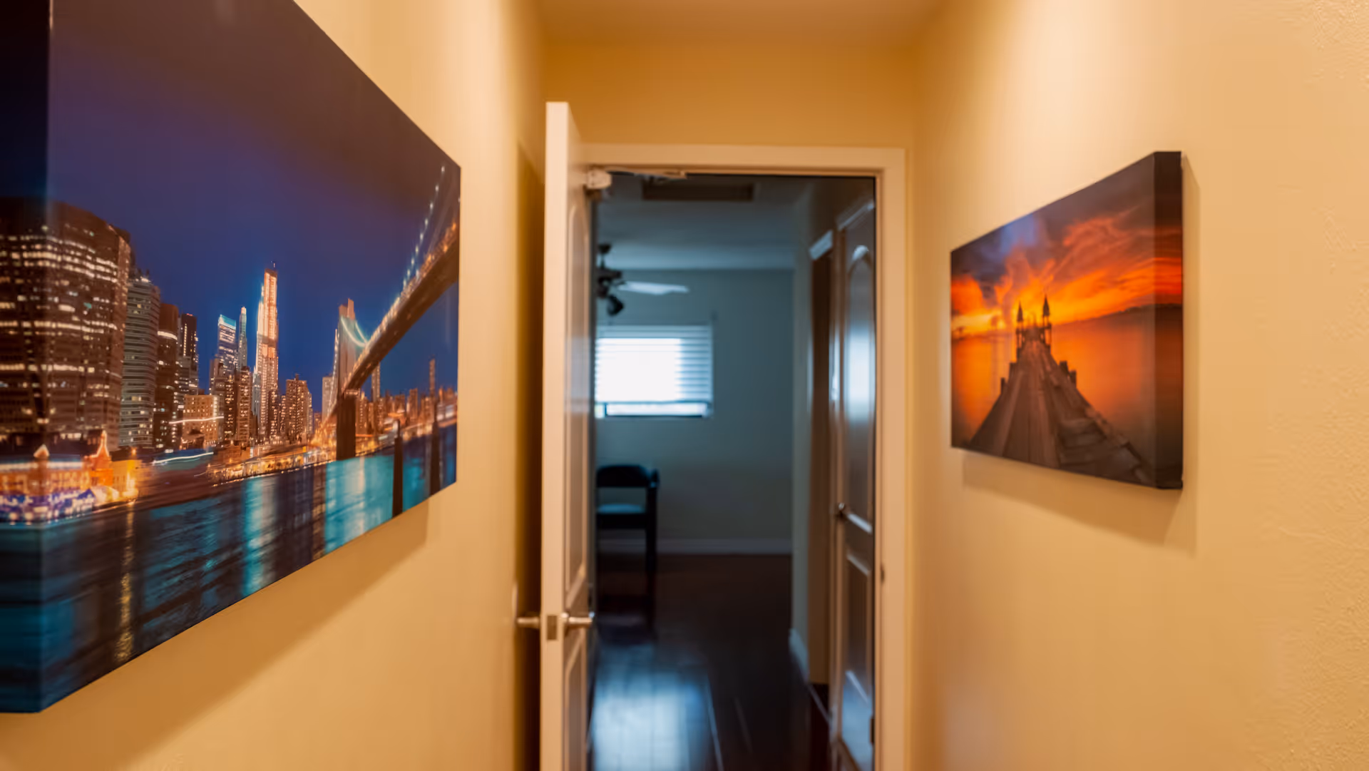 A narrow interior hallway with framed artwork on both walls leading to an open door and a room beyond.