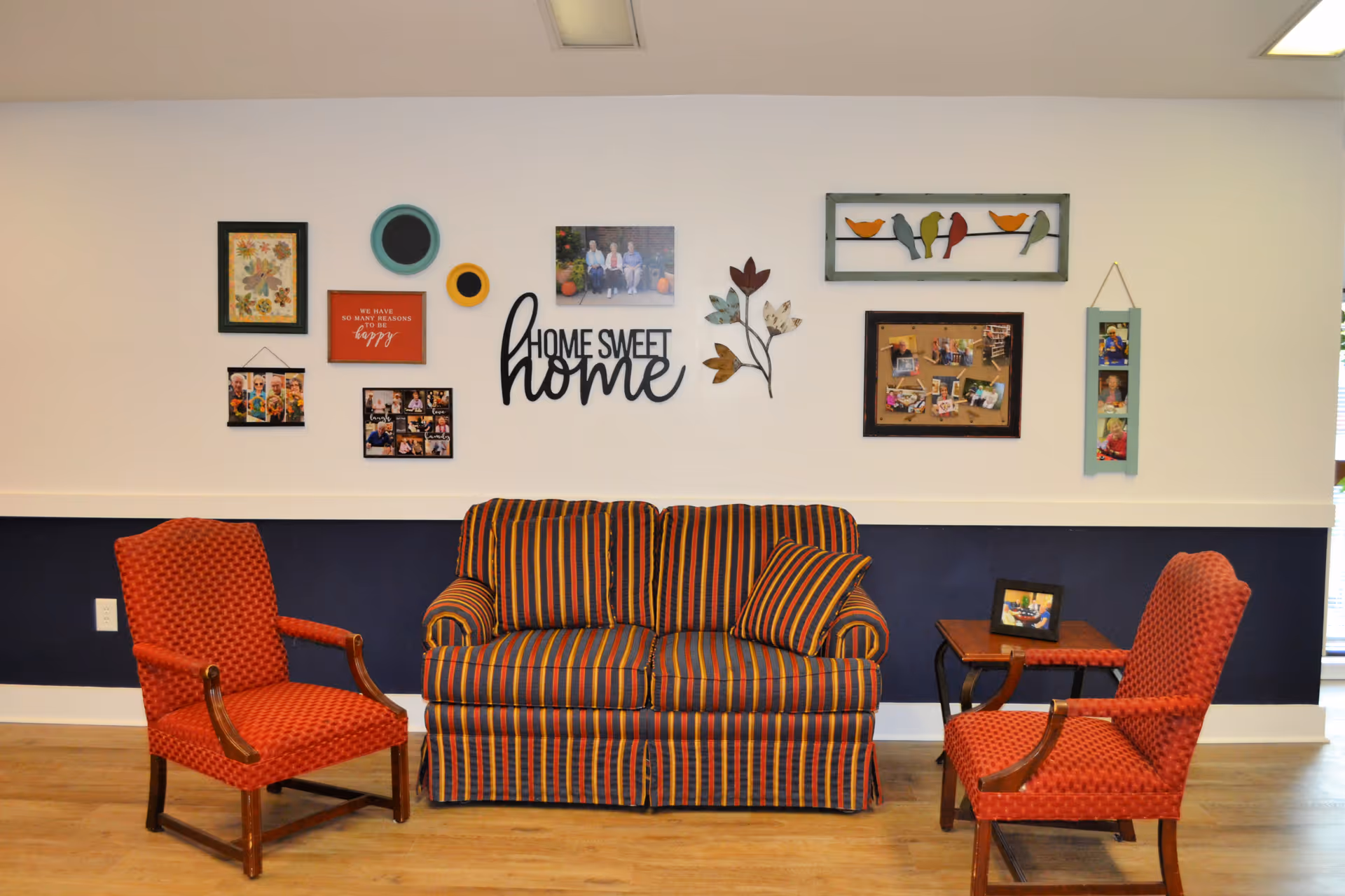 A cozy sitting area with a striped loveseat and two red patterned armchairs facing each other. The wall behind features various framed photos, decorative plates, a metal wall art of birds on a wire, and a sign that says 'Home Sweet Home'. The lower half of the wall is painted dark blue, and the floor is wooden.