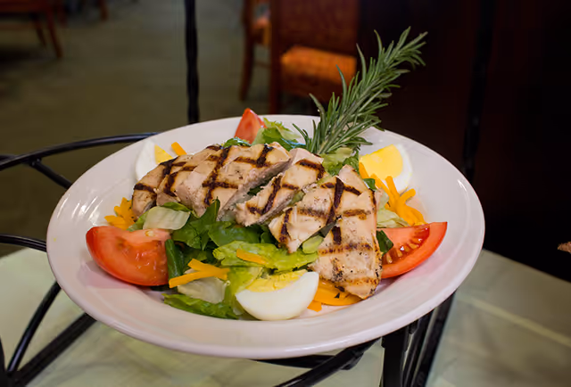 Grilled chicken salad with sliced tomatoes, hard‑boiled egg, shredded cheese and a rosemary garnish on a white plate.