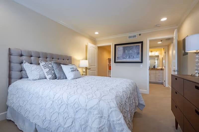 A neatly made bed with a tufted headboard and decorative pillows in a bedroom. There is a bedside table with a lamp on the left side of the bed. On the right side of the room, there is a wooden dresser with a lamp on top. The room has beige walls and carpeted floor. A doorway leads to a bathroom with a vanity and mirror visible.