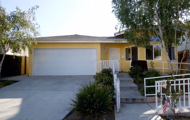 Front exterior view of a single-story yellow residential building with a white garage door, a small porch with steps and white railings, surrounded by greenery and trees.