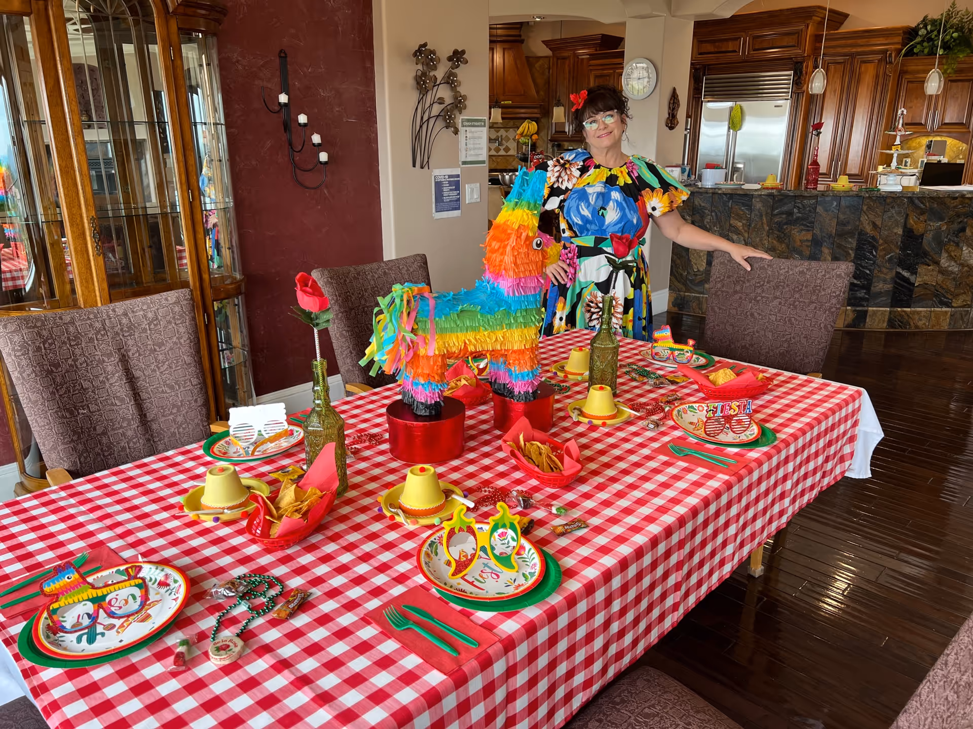A woman in a colorful floral dress stands next to a dining table covered with a red and white checkered tablecloth. The table is decorated with festive Mexican-themed items including a colorful piñata centerpiece, sombrero-shaped cups, novelty glasses, and plates with chili pepper designs. The background shows a kitchen area with wooden cabinets and stainless steel appliances.