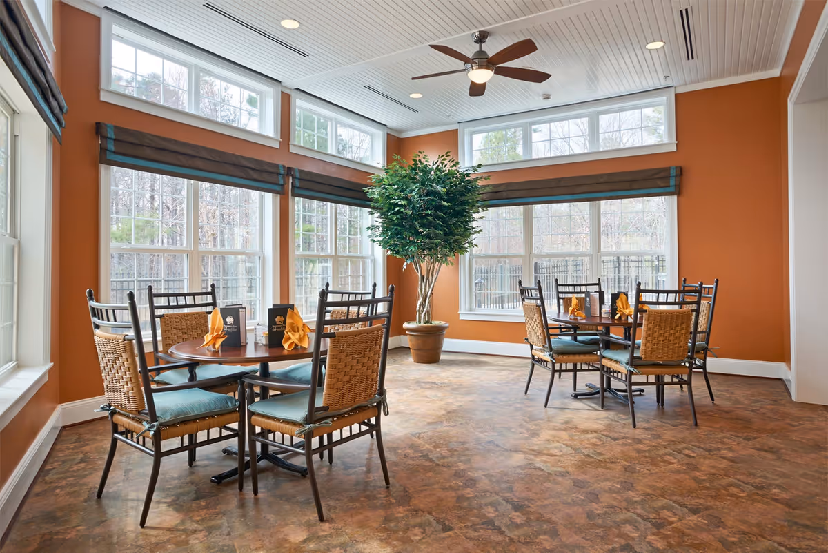 Sunlit dining room with round wooden tables, wicker-backed chairs, folded napkins, and large windows with a potted tree.