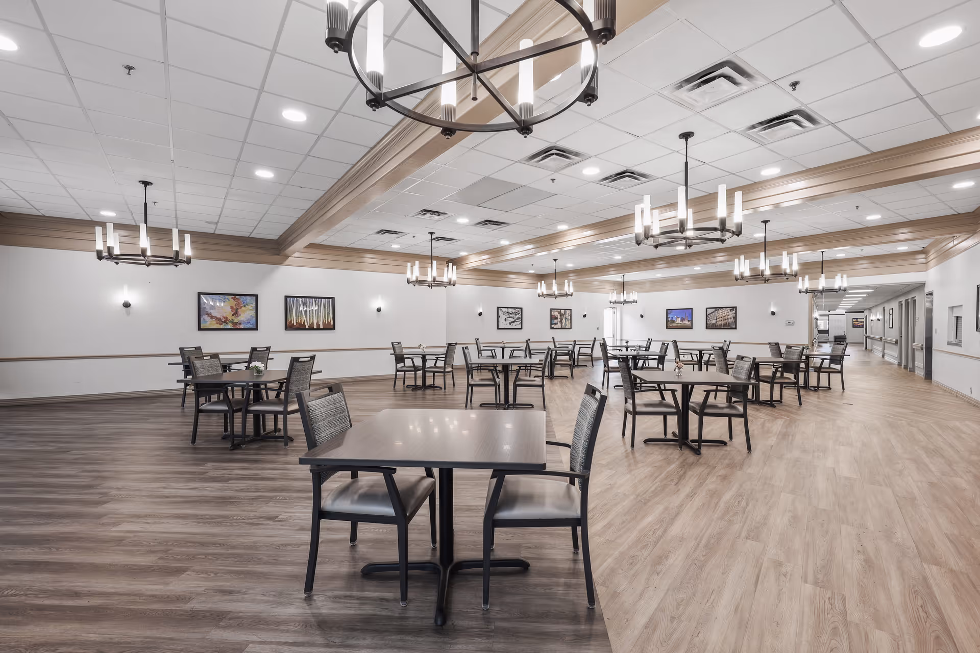 A spacious dining room with multiple square tables and chairs arranged neatly. The room features wood flooring, white walls with framed artwork, and several modern chandeliers hanging from a ceiling with recessed lighting and wooden beams.