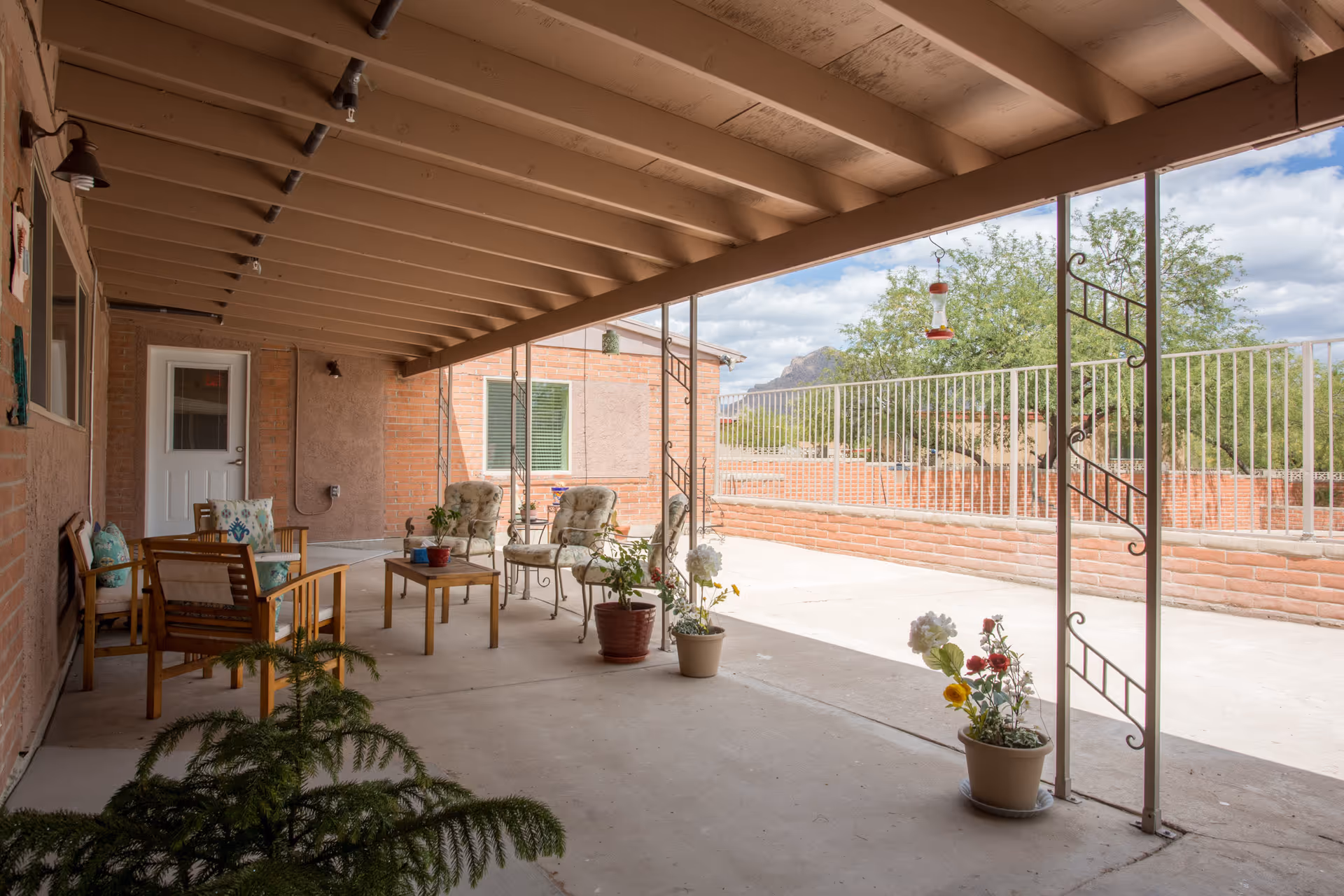 Covered outdoor patio area with wooden ceiling beams, several chairs with cushions, a small table, and potted plants. The patio overlooks a fenced yard with trees and a partly cloudy sky.
