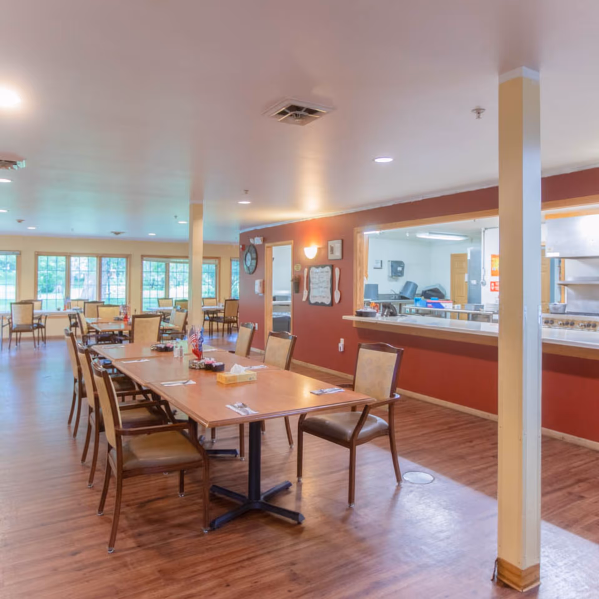 A spacious dining room in a senior living facility with wooden floors, multiple tables and chairs arranged neatly, large windows letting in natural light, and a serving window to the kitchen area with red walls.