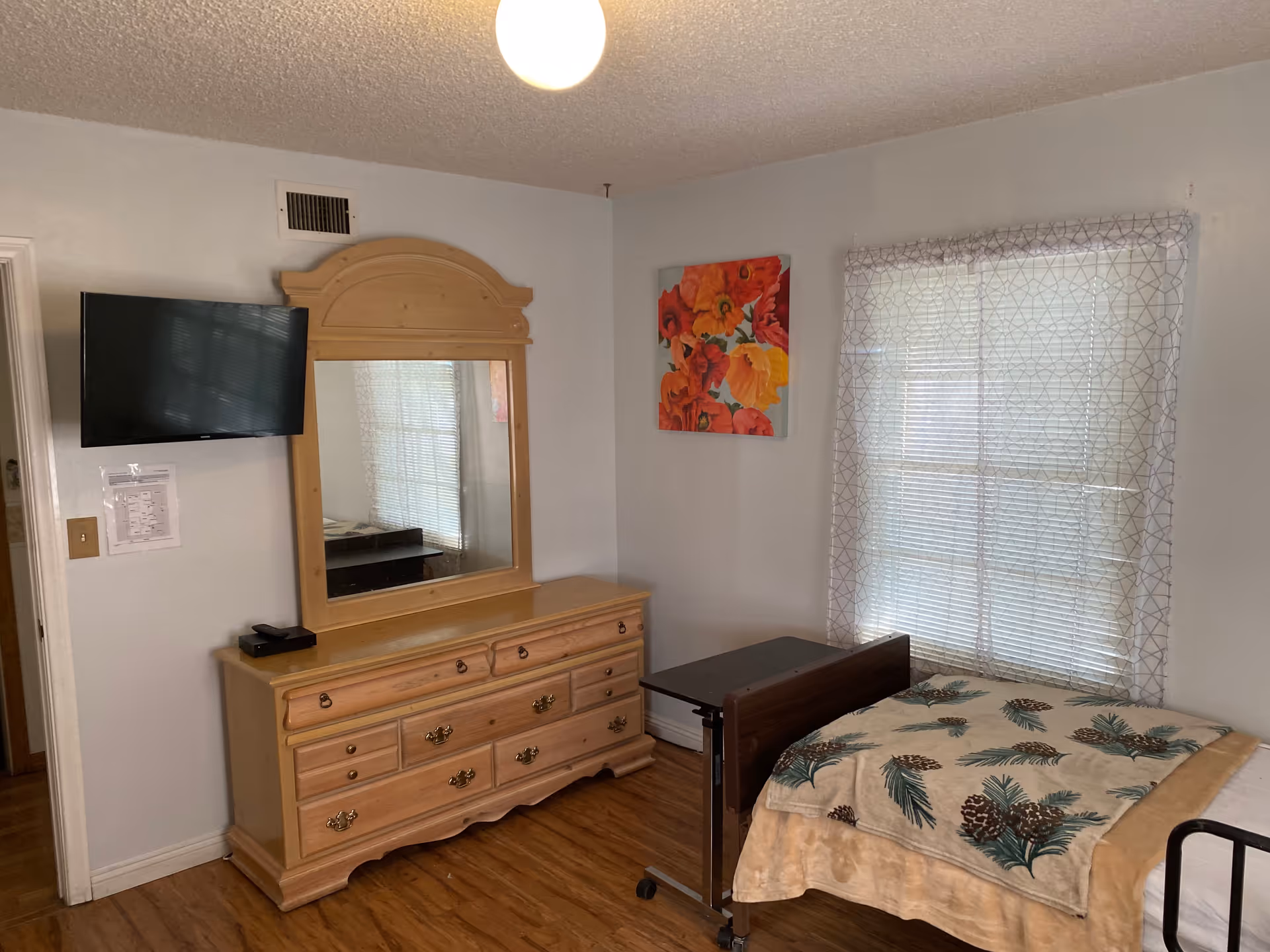 A bedroom with a wooden dresser and mirror, a mounted flat-screen TV on the wall, a window with sheer patterned curtains, a bed with a pinecone and leaf patterned blanket, and a small overbed table.