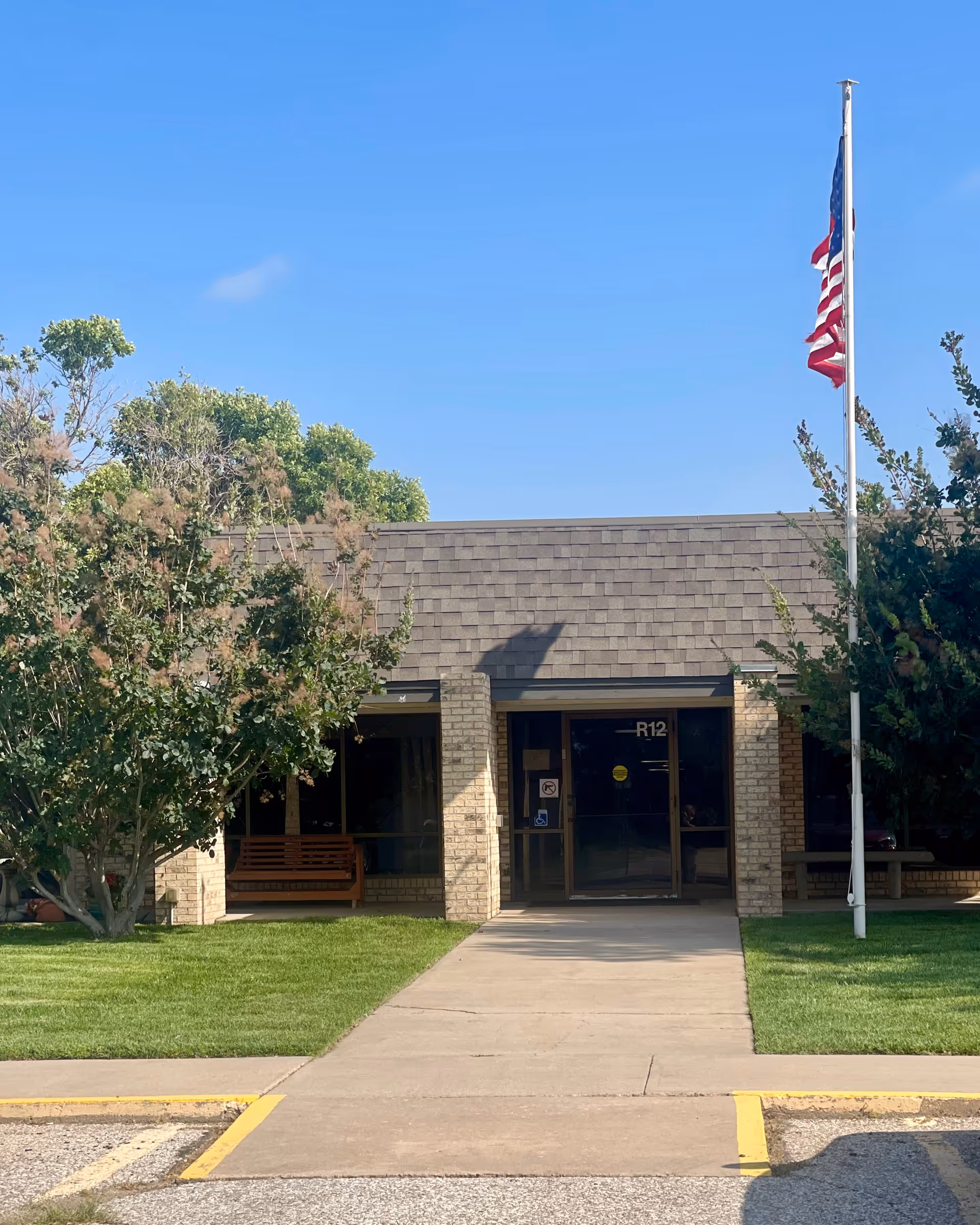 Front entrance of a single-story brick building with a shingled roof, flanked by green trees and a neatly maintained lawn. A concrete walkway leads to glass double doors marked with 'R12'. An American flag on a flagpole is visible to the right of the entrance under a clear blue sky.
