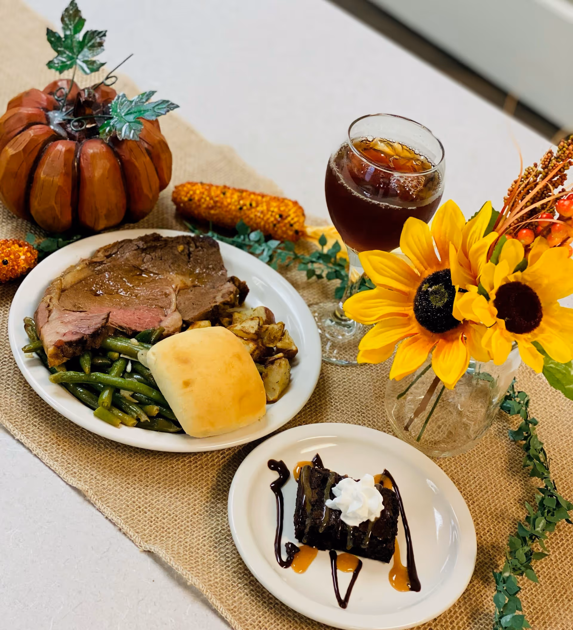 A meal served on a table with a plate containing a slice of roast beef, green beans, roasted potatoes, and a dinner roll. Next to it is a small plate with a chocolate dessert topped with whipped cream and drizzled with caramel and chocolate sauce. A glass of iced tea and a vase with bright yellow sunflowers are also on the table, along with decorative autumn-themed items including a small pumpkin and corn cobs.