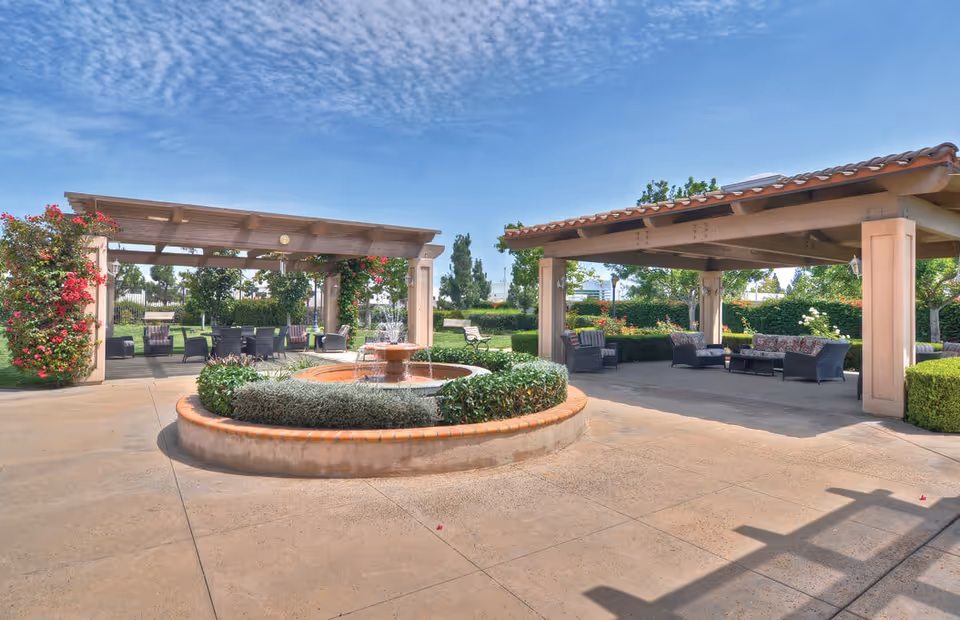 Outdoor patio area at Ivy Park at Cerritos featuring two covered seating areas with wicker furniture and cushions, a central circular fountain surrounded by greenery, and a clear blue sky with scattered clouds.