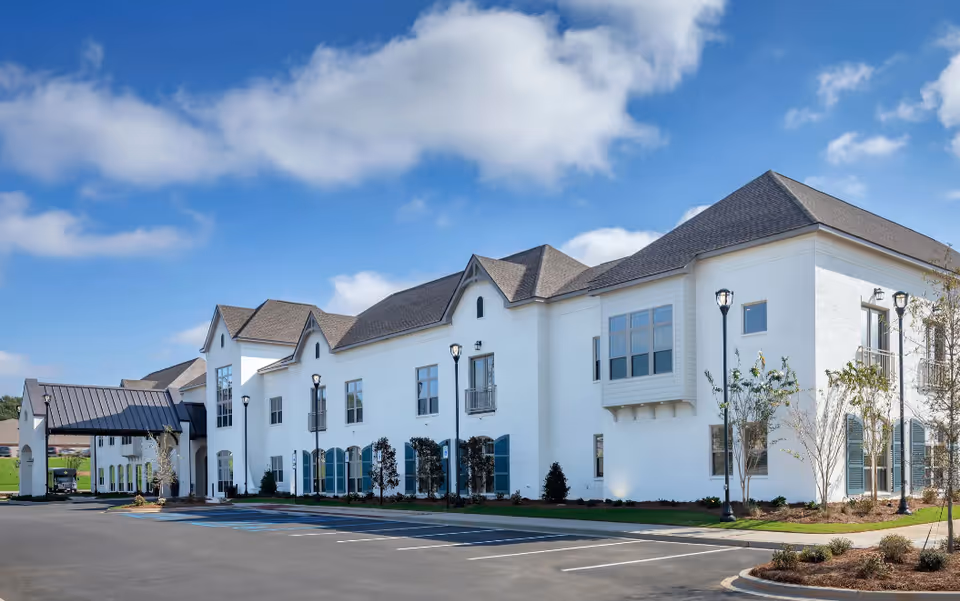 White two-story building with gabled roofs and a covered entrance beside a parking lot under a blue sky.