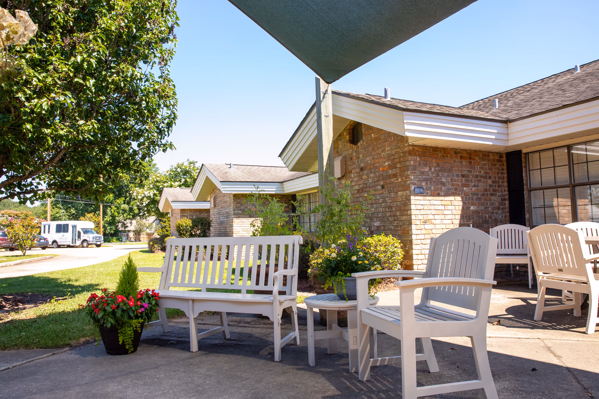 Outdoor seating area at Lexington House with white benches and chairs arranged around small tables on a concrete patio. There are potted plants with flowers and greenery, a large tree providing shade, and brick buildings in the background under a clear blue sky.