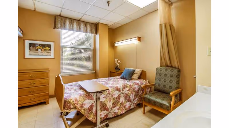 A nursing facility bedroom with a single bed covered in a floral quilt, a small overbed table, a green patterned armchair, a wooden dresser, a window with blinds and a valance, and a wall-mounted light above the bed.