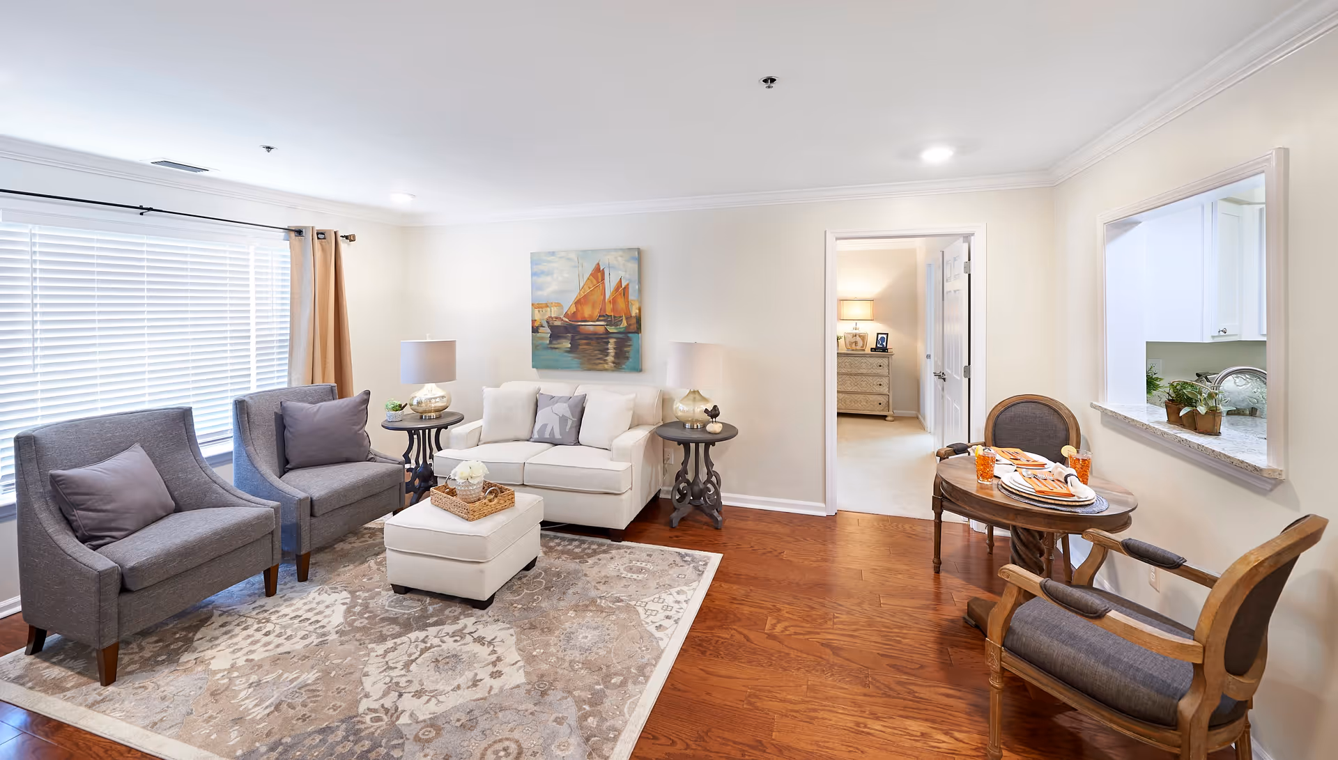 A bright and cozy living room with two gray armchairs and a white loveseat arranged around a patterned area rug. There are two side tables with lamps on either side of the loveseat. A painting of sailboats hangs on the wall above the loveseat. To the right, there is a small round dining table set for two with wooden chairs, and a pass-through window to the kitchen. Hardwood floors and large windows with blinds and curtains complete the room.