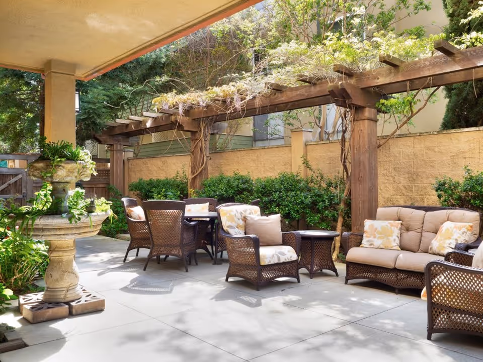 Outdoor patio area with wicker furniture including a sofa, armchairs, and a table under a wooden pergola with climbing plants. There is a stone fountain on the left side and greenery along the back wall.