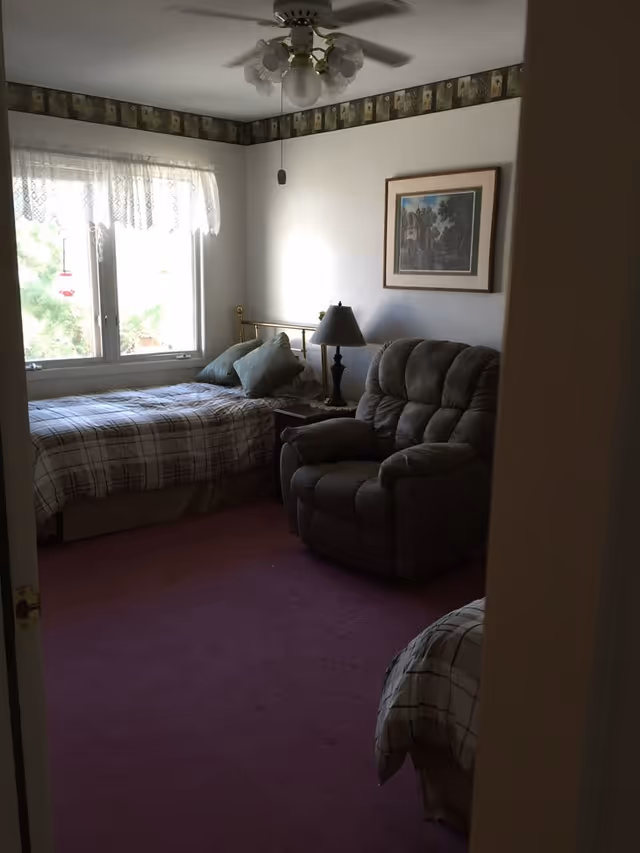 Cozy bedroom with two twin beds, a plush recliner, bedside lamp, ceiling fan, and a window with lace curtains.