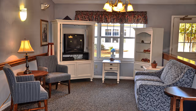 Cozy common-area living room with upholstered chairs and a patterned sofa, a TV in a white cabinet, side tables, and a window letting in daylight.