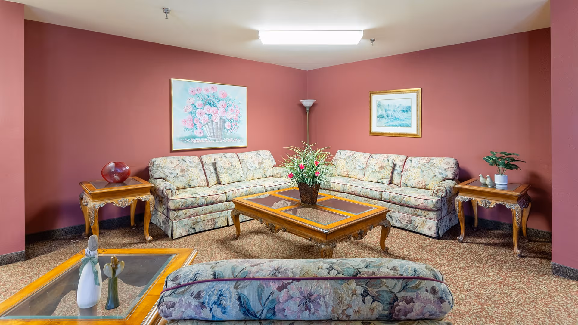 A cozy living room with floral patterned sofas arranged around a wooden coffee table with a glass top. The room has red walls adorned with two framed paintings, a floor lamp in the corner, and two wooden side tables with decorative items including a red glass bowl and a small plant. The carpet has a floral pattern complementing the furniture.