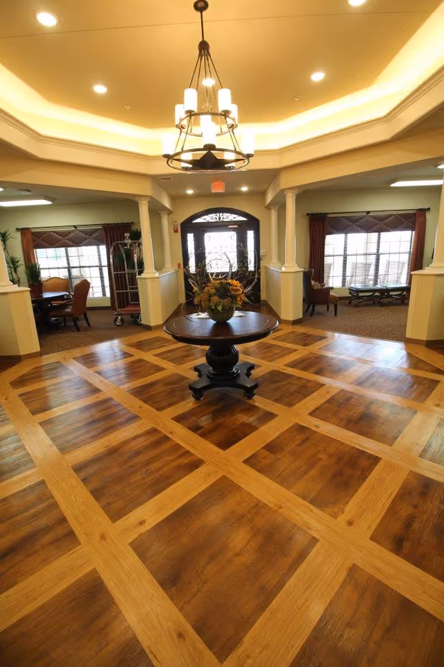 Interior view of a senior living facility lobby with a decorative wooden floor featuring a grid pattern. In the center, there is a round wooden table with a floral arrangement. The ceiling has recessed lighting and a chandelier. The background shows large windows with curtains and seating areas on both sides, along with a glass entrance door.