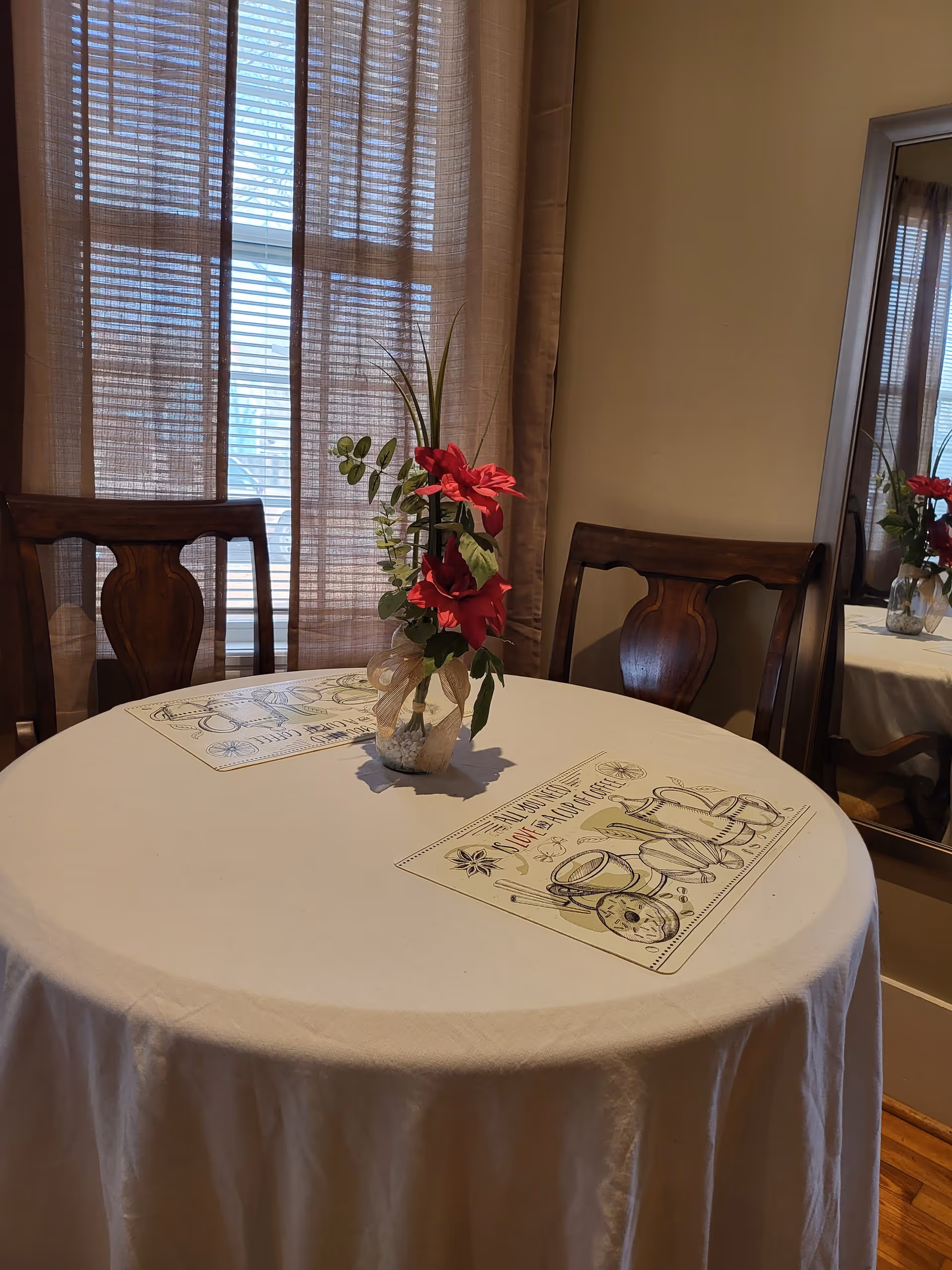 A round dining table covered with a white tablecloth, set with two placemats featuring kitchen-themed illustrations and text. A small vase with red flowers and green leaves is placed at the center of the table. Two wooden chairs with carved backs are positioned behind the table. Sheer curtains cover a window in the background, and a large mirror on the right side reflects part of the table and flowers.