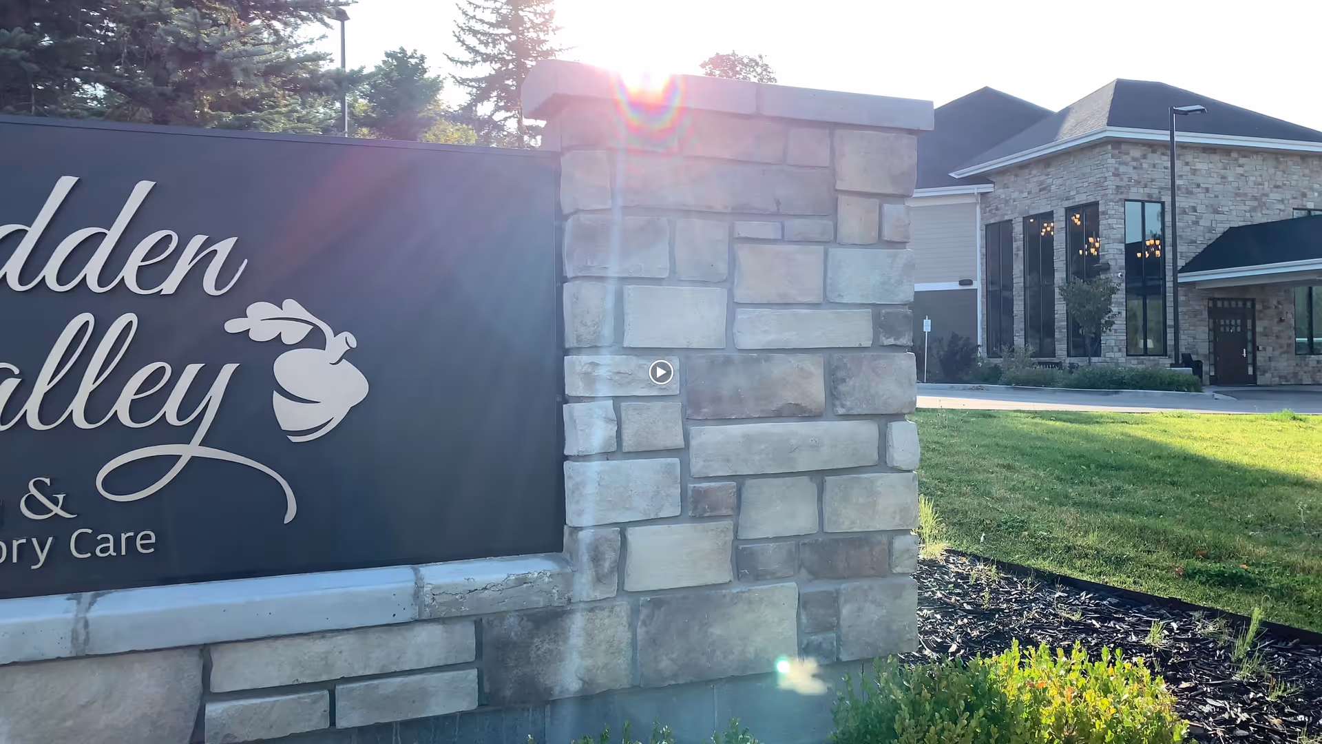 Stone and concrete signpost with a black sign displaying the text 'Hidden Valley | Assisted Living and Memory Care' next to a grassy area and a building with large windows in the background under sunlight.