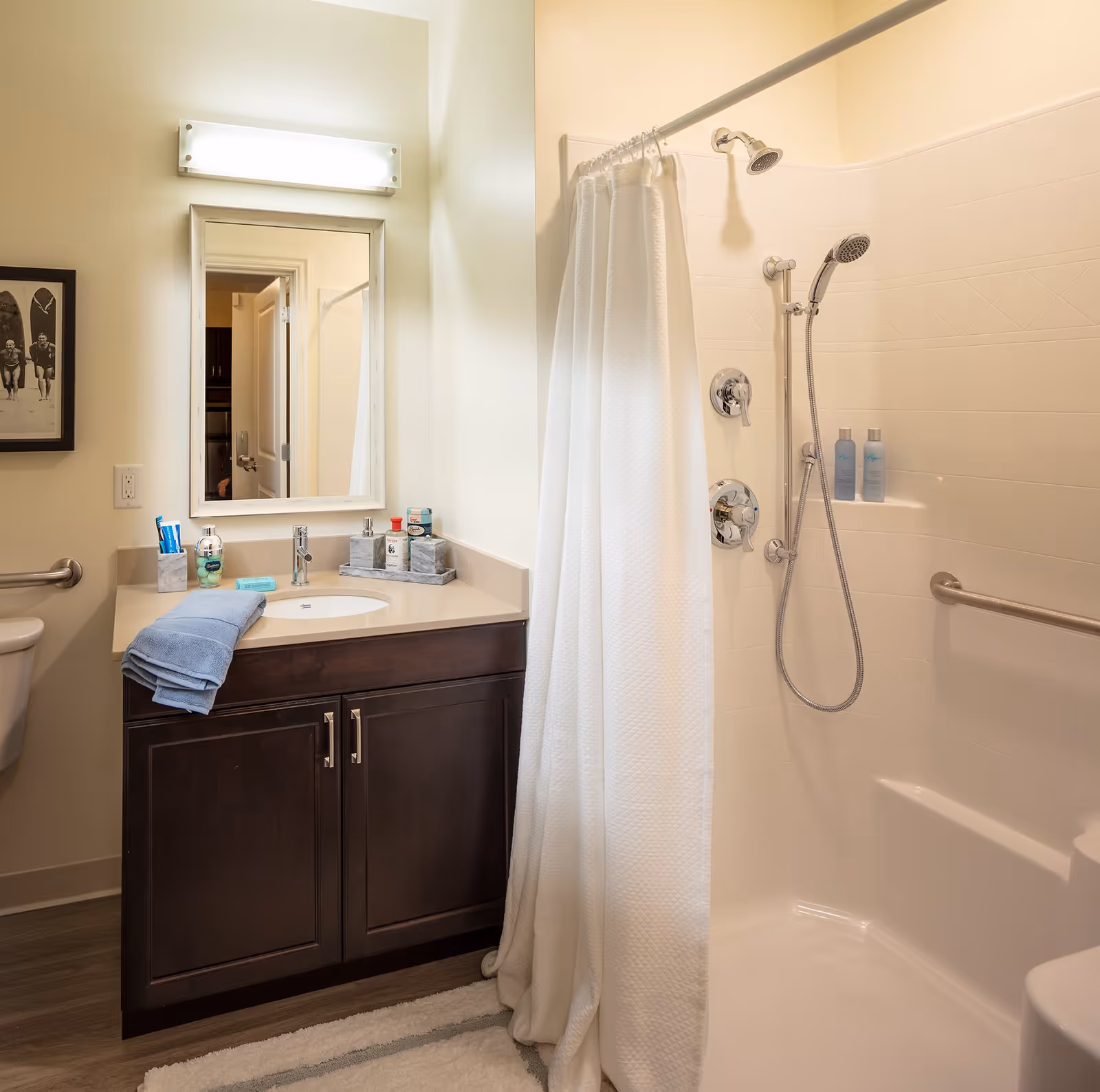 A clean and well-lit bathroom featuring a dark wood vanity with a beige countertop, a sink, a mirror with a light fixture above it, a white shower curtain partially drawn to reveal a shower area with a handheld and fixed showerhead, grab bars, and toiletries on a built-in shelf. A blue towel is folded on the countertop and a framed black and white photo is on the wall.