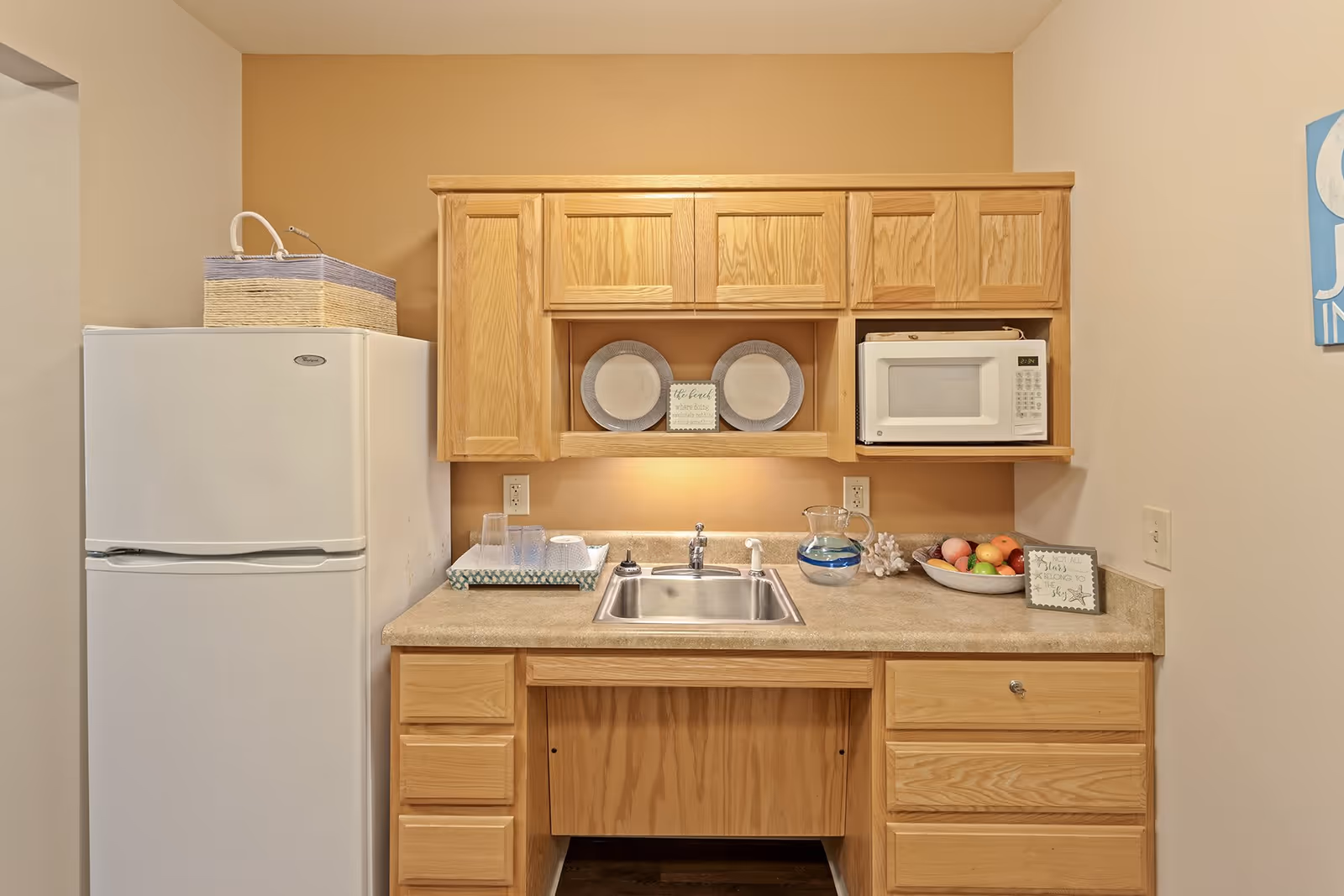 A small kitchen area with light wood cabinets, a white refrigerator on the left, a stainless steel sink in the center, and a white microwave on the right. The countertop has a tray with cups, a glass pitcher with water, a bowl of fruit, and decorative signs and plates on the wall and countertop.