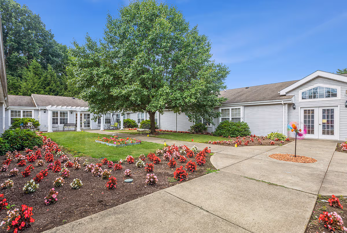Outdoor courtyard area at The Addison of Lowrie Place featuring a large tree in the center, surrounded by flower beds with red and pink flowers, green grass, and a paved walkway. The building with white siding and multiple windows surrounds the courtyard under a clear blue sky.