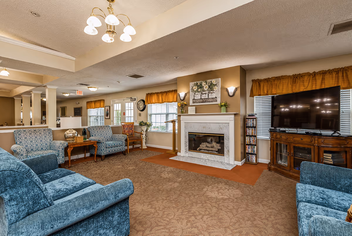 A cozy living room area in a senior living facility with blue upholstered armchairs and sofas arranged around a white marble fireplace. Above the fireplace is a floral painting and two wall sconces. To the right, there is a large flat-screen TV on a wooden cabinet with DVDs stacked beside it. The room has beige walls, brown carpet, and windows with orange valances letting in natural light. A chandelier hangs from the ceiling, and there are additional chairs and tables near the windows.